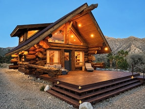 A log cabin with a wooden deck, a front porch, and a mountain backdrop.