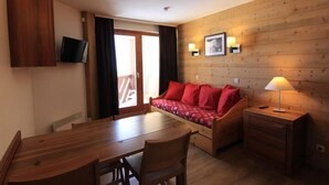 Ein Zimmer mit Holzvertäfelung, einem Esstisch, einer Couch mit roten Kissen, einem an der Wand montierten Fernseher und einem Nachttisch mit Lampe.
