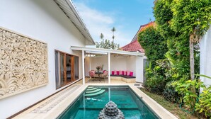 Una piscina con una scultura decorativa in primo piano, una zona patio coperta con posti a sedere e una rigogliosa vegetazione intorno alla proprietà.