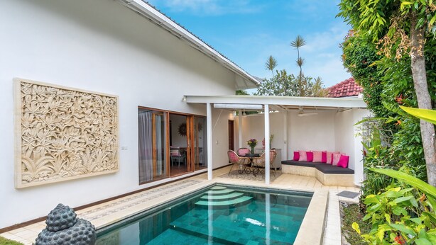 Una piscina con acqua cristallina blu, circondata da un muro bianco con pannello decorativo, una zona relax coperta con cuscini e una statua in primo piano.