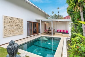 Une piscine à l’eau d’un bleu limpide, entourée d’un mur blanc orné d’un panneau décoratif, un espace de détente couvert avec des coussins, et une statue au premier plan.