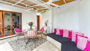 Un'accogliente area patio con una porta vetrata che dà su un balcone, mobili in vimini e un divano nero con cuscini rosa.