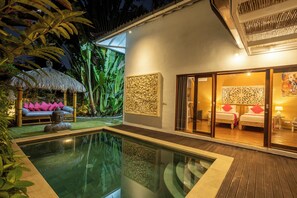 Eine moderne Villa mit Swimmingpool, einer Sitzgelegenheit im Freien unter einem Reetdach und einem Schlafzimmer mit großem Fenster.