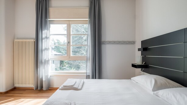 Un dormitorio moderno con una cama grande, una ventana con cortinas, un radiador y un cabecero con compartimentos de almacenamiento.