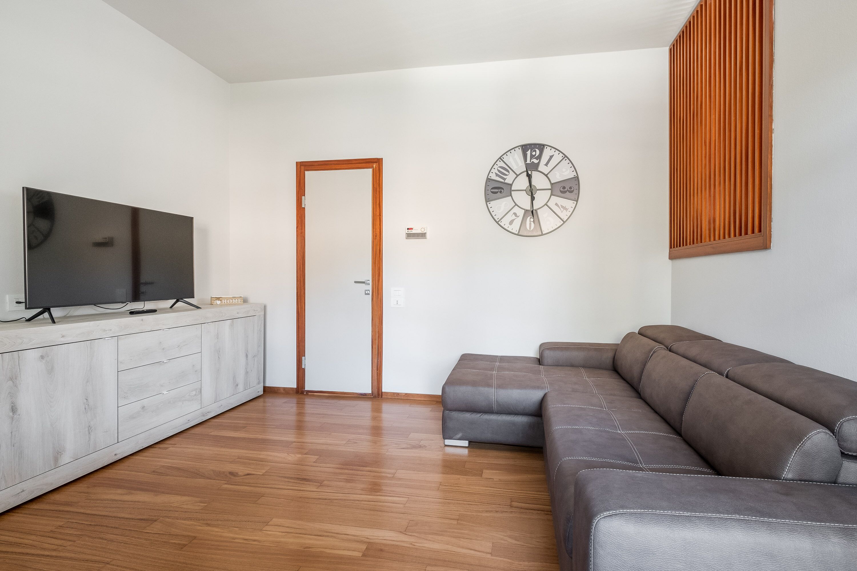 Ein modernes Wohnzimmer mit einem grauen Ecksofa, einem Flachbildfernseher auf einer weißen Kommode, einer Holztür und einer Wanduhr.