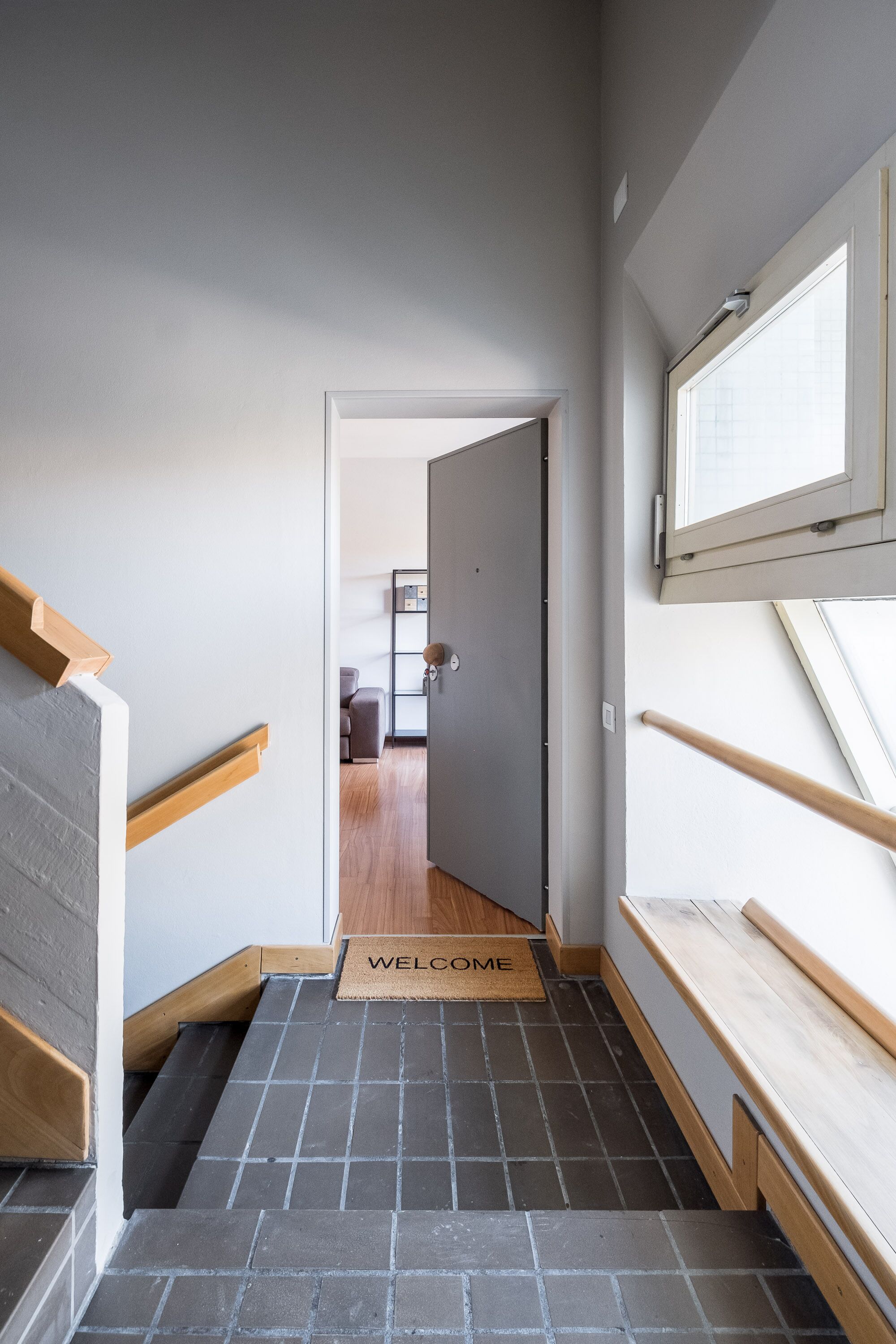 A staircase with a welcome mat leading to a room with a wooden floor and a window.