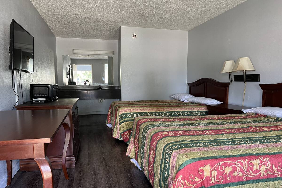 A hotel room with a wooden desk, two beds with patterned bedding, a television, and a window.