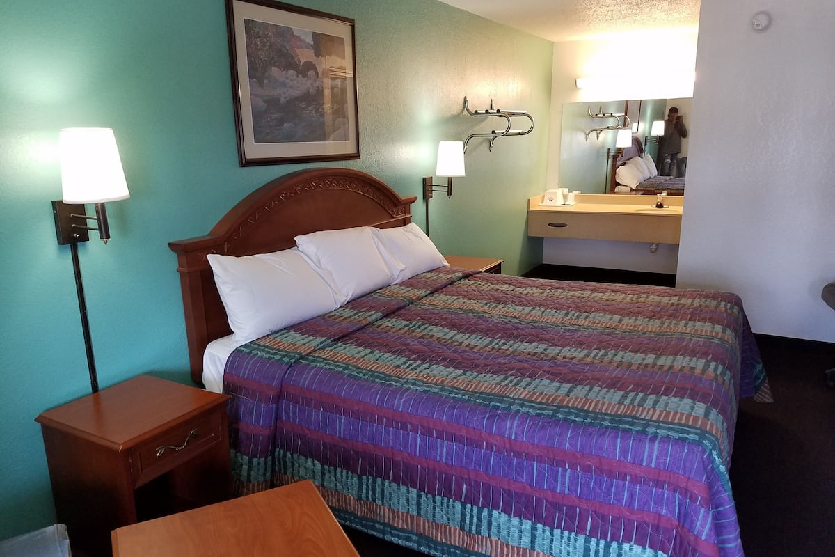 A hotel room with a bed, bedside lamps, a wooden nightstand, a small table, a mirror, and a framed picture on the wall.