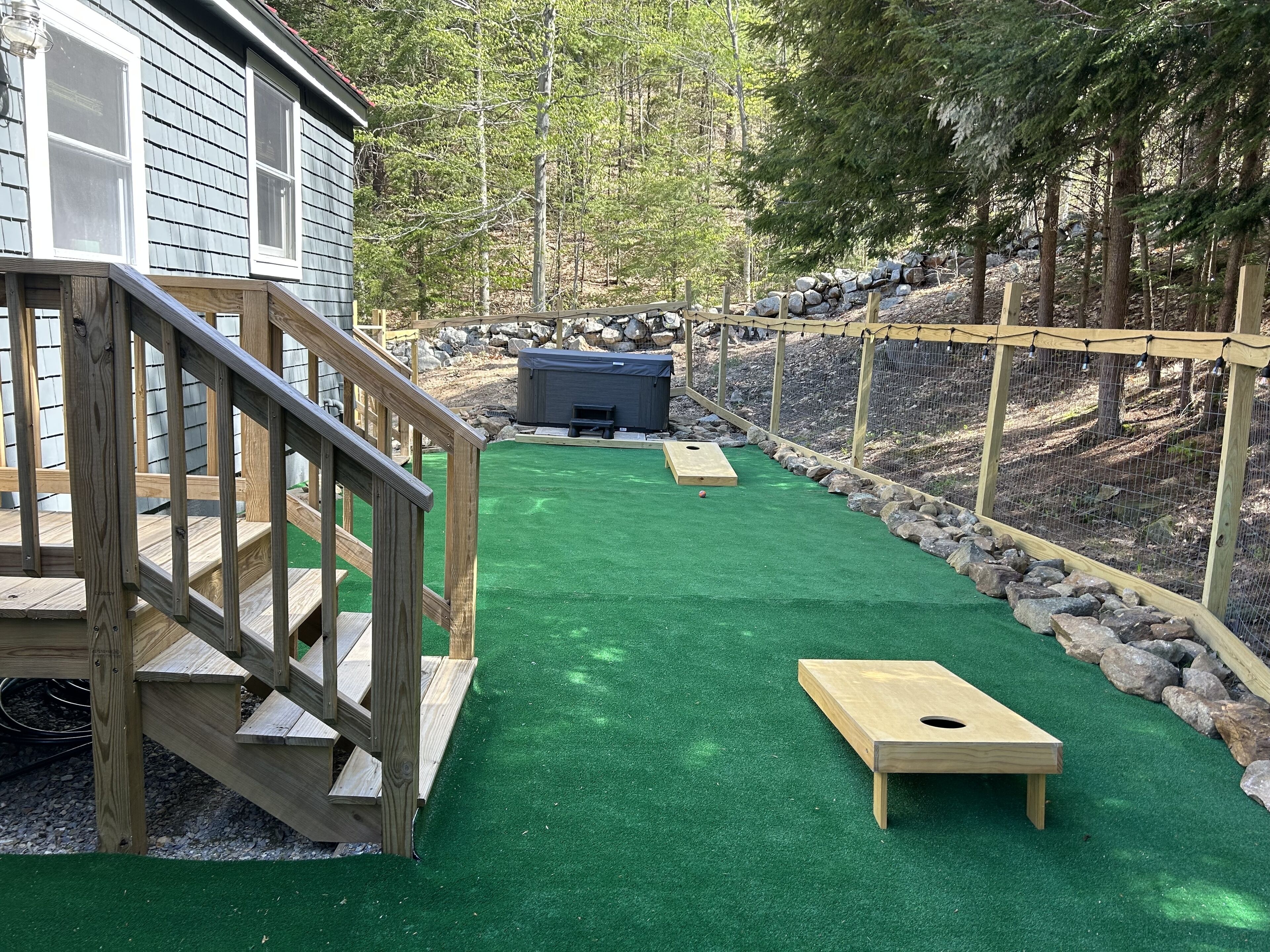 A wooden deck with a ramp leading to a house, a green artificial turf area with a wooden target, and a stone wall.