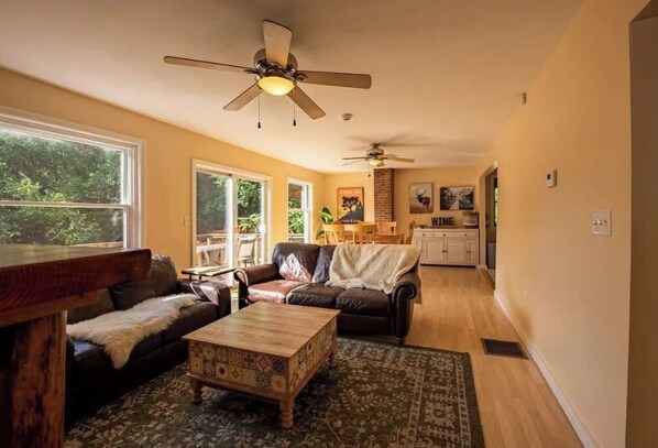 A spacious living room with a large window, a ceiling fan, a wooden coffee table, and a patterned rug.
