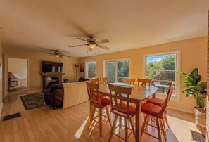 A spacious living room with a wooden dining table and chairs, a fireplace, and large windows offering a view of the outdoors.
