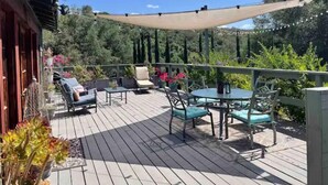 Una terraza de madera con mobiliario exterior, incluyendo sillas y una mesa, rodeada de follaje y árboles.