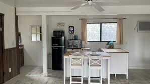 Una cocina con refrigerador, microondas y tostadora, un comedor con mesa y sillas, un ventilador de techo y una ventana con cortinas.