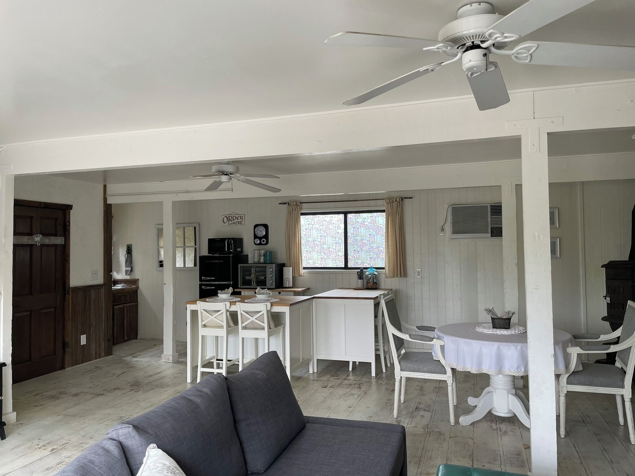 A spacious living area with a grey sofa, a dining table with white chairs, a kitchen area with appliances, and a ceiling fan.