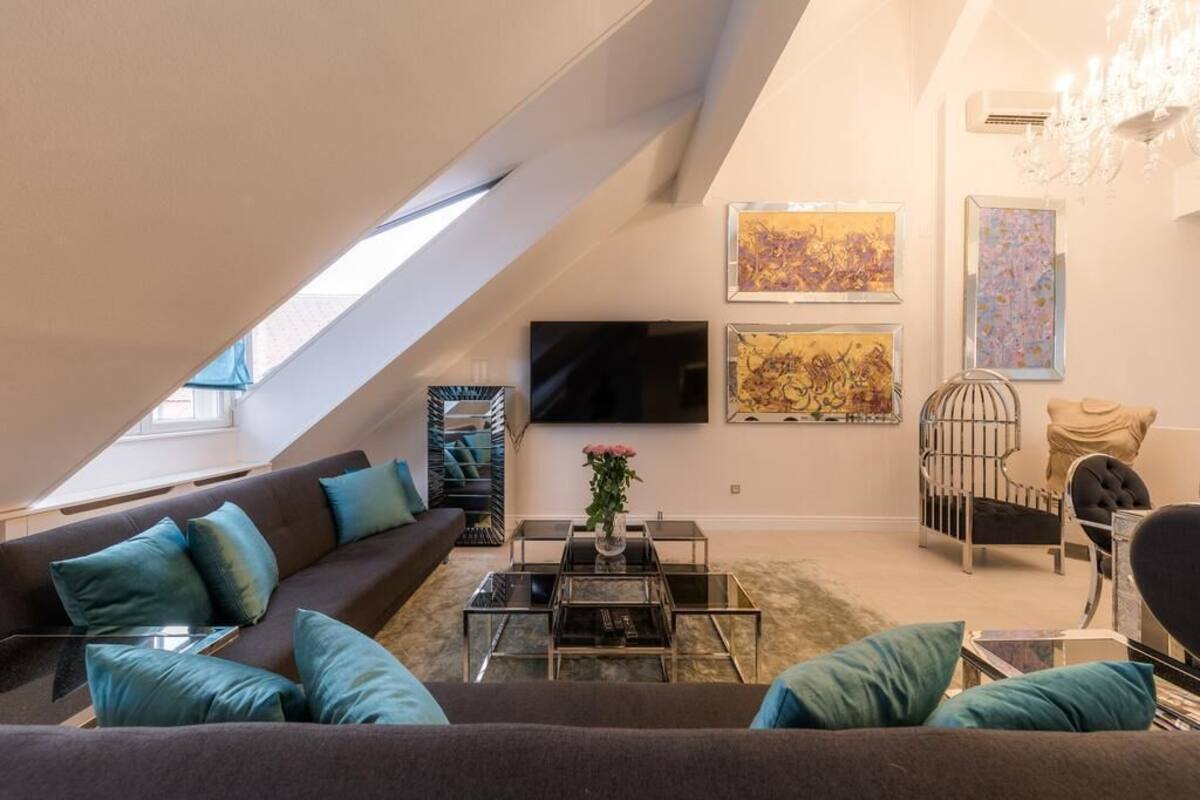 Ein modernes Wohnzimmer mit schräger Decke, einem dunklen Sofa mit türkisfarbenen Kissen und einem Glastisch. An den Wänden befindet sich Kunstwerk.