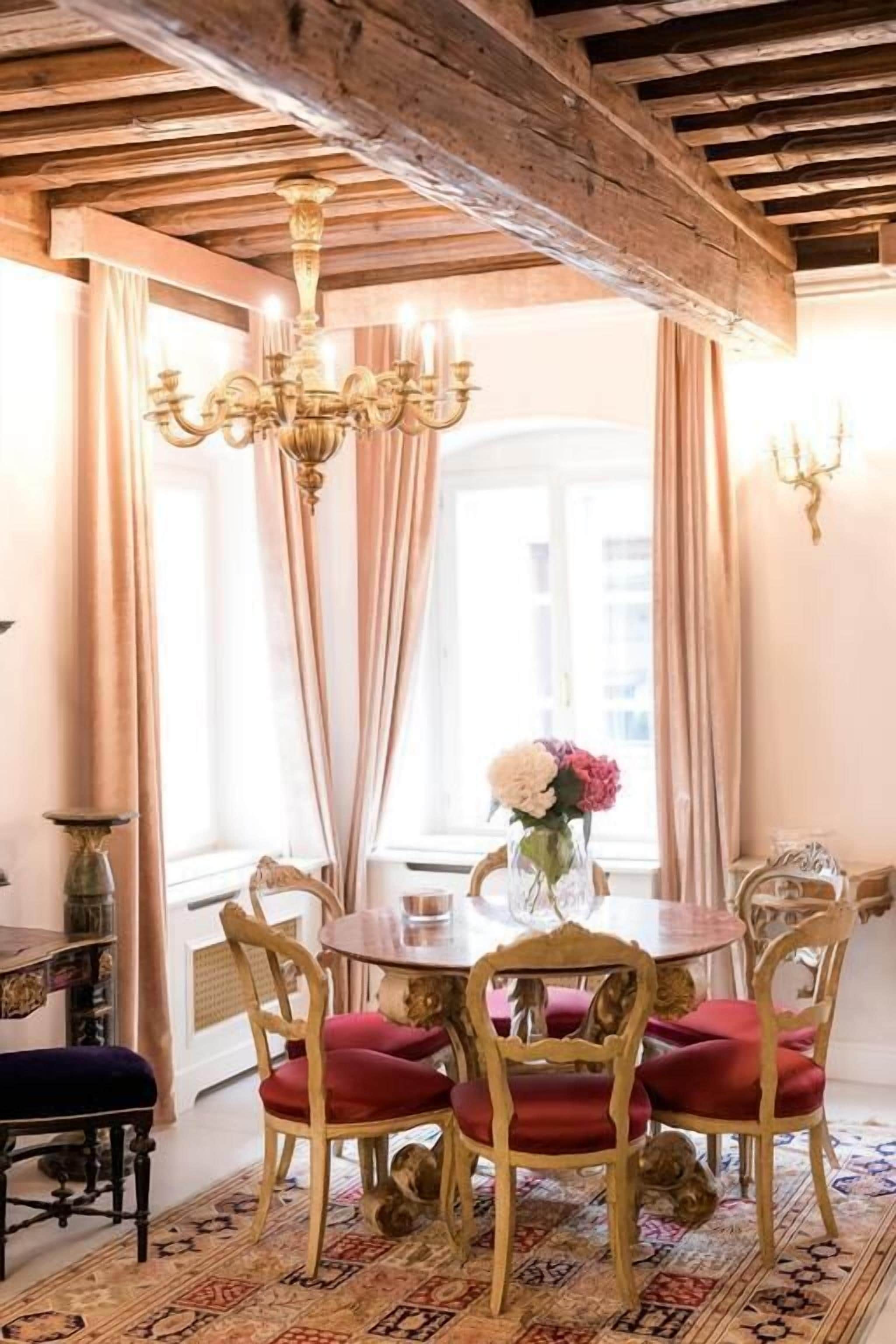 A dining room with a round table, six chairs, a chandelier, and a window with curtains.