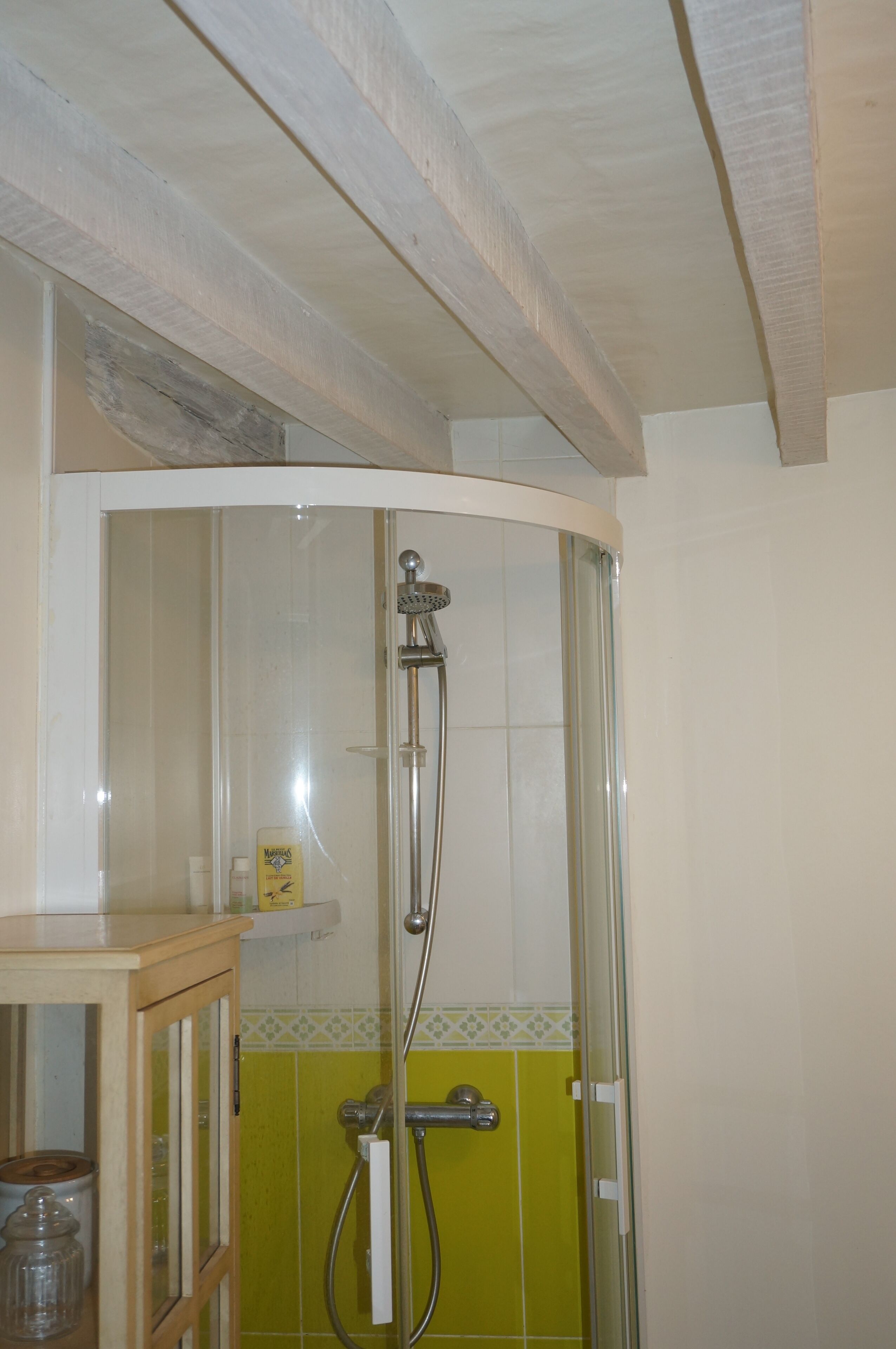 Ducha con mampara de vidrio, mueble de madera con frascos y pared revestida de azulejos con detalle amarillo.