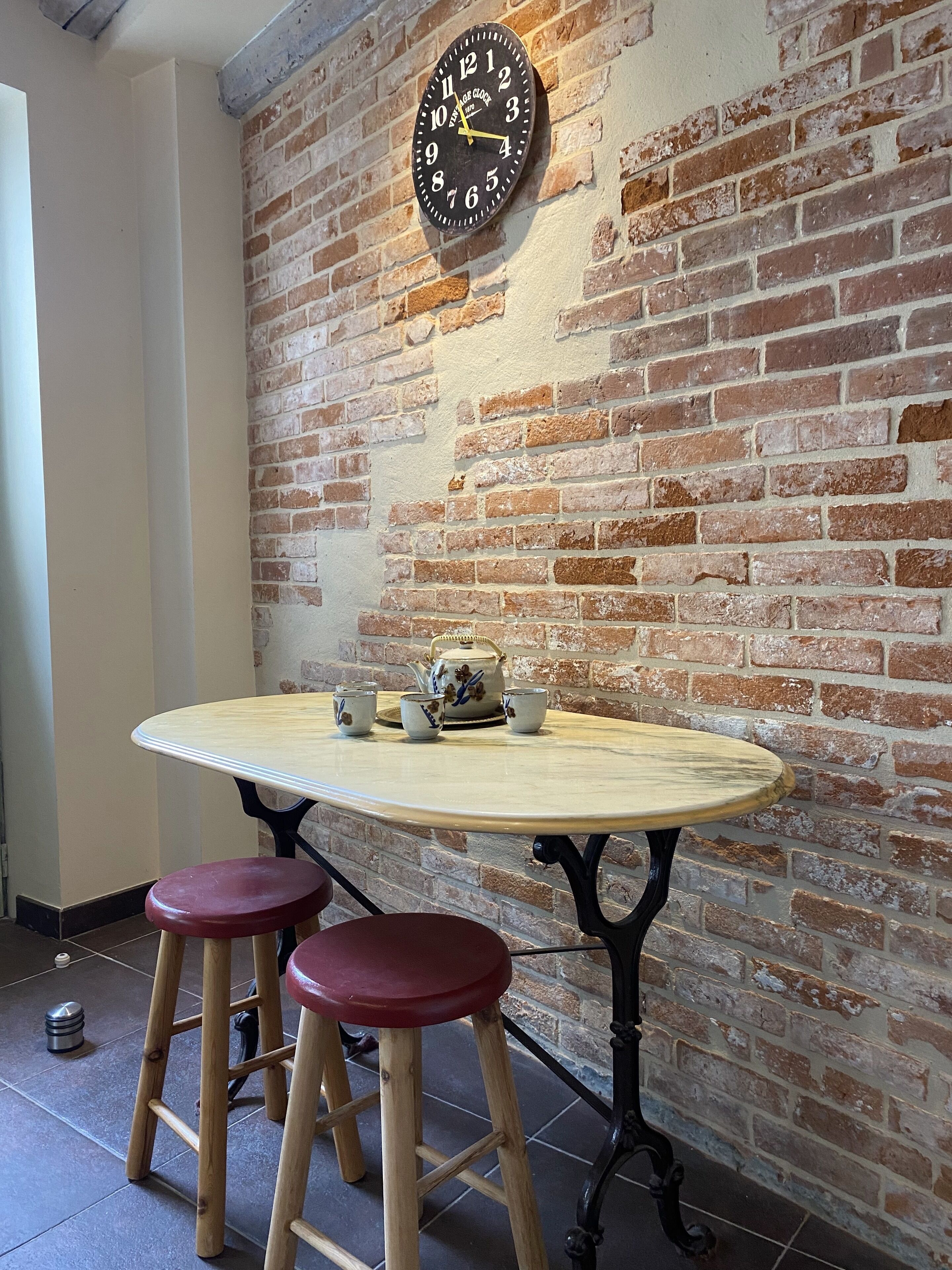 A round table with stools, a clock, and a teapot on a brick wall.