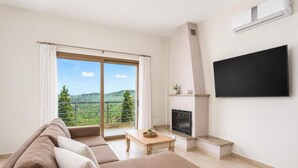 A living room with a sofa, a fireplace, a television, and a balcony with a view of greenery.