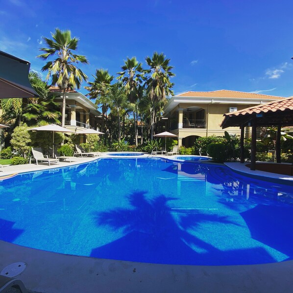 A swimming pool with lounge chairs and umbrellas, surrounded by palm trees and a building with a tiled roof.