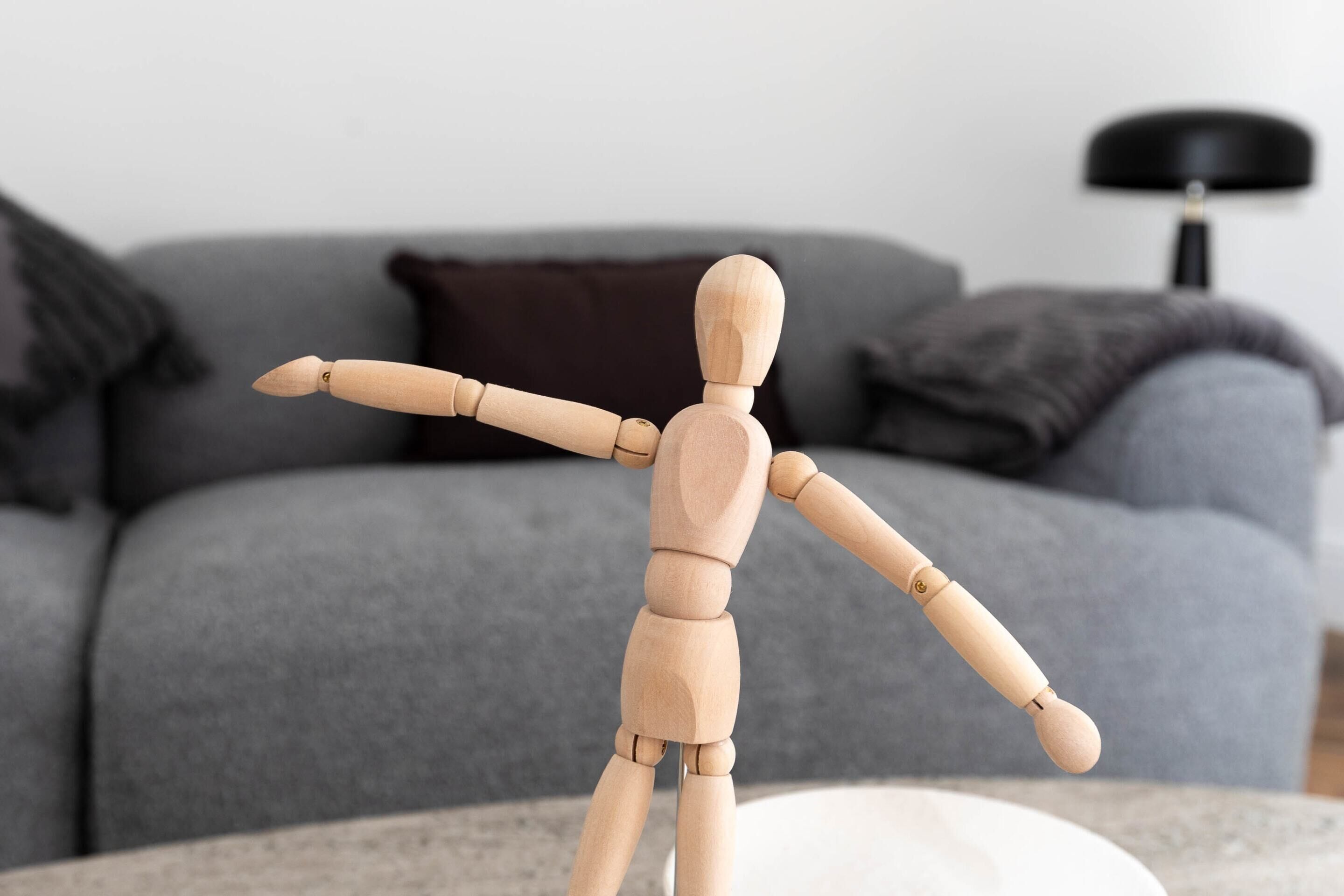 A wooden mannequin in a living room with a grey sofa and a black lamp in the background.