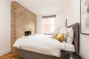 A bedroom with a brick wall, a bed with white bedding, a gold lamp, and a window with a view of a brick building. - Stylish 2 Bedroom Apartment Near Oxford Street (London)