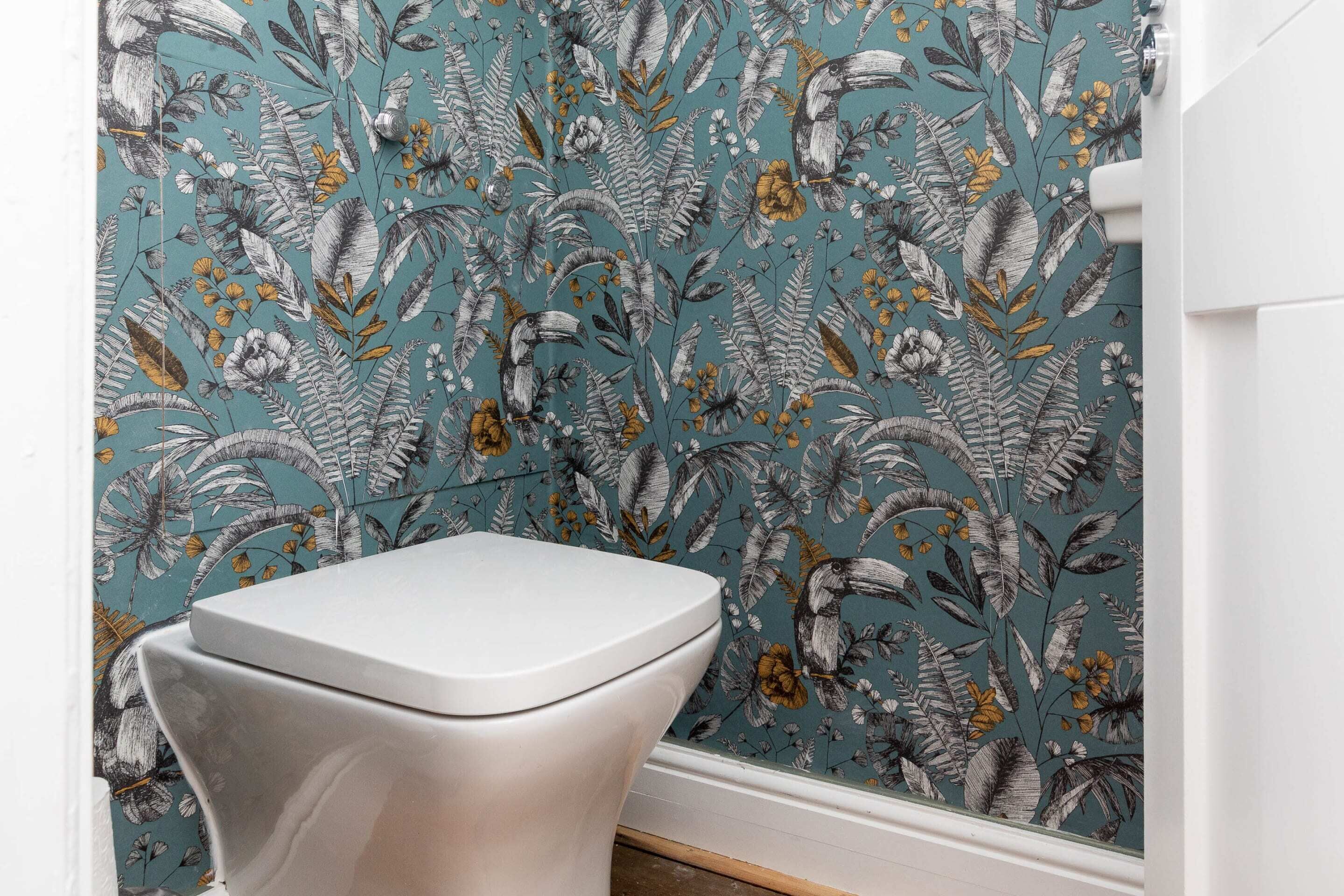A bathroom with a toilet, a white wall, and a floral wallpaper pattern.
