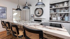 Une cuisine moderne dotée d’un plan de travail en marbre, de chaises noires, de luminaires suspendus et d’une étagère où sont exposés divers ustensiles de cuisine.