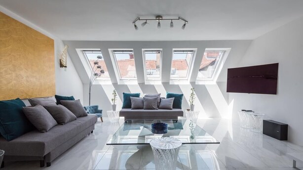 Ein modernes Wohnzimmer mit einem Glastisch, einer Couch und einem an der Wand montierten Fernseher. Dachfenster sorgen für natürliches Licht im Raum.