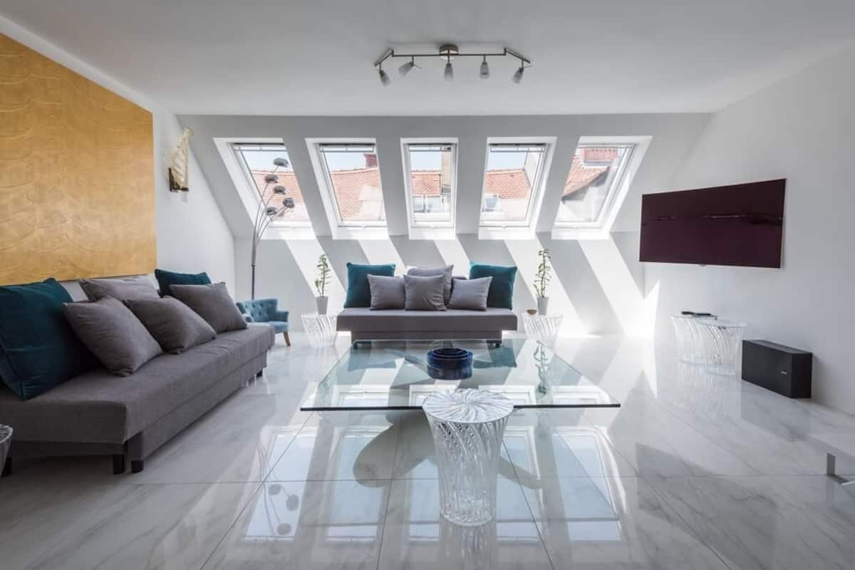 Una sala de estar moderna con una mesa de centro de vidrio, un sofá y un televisor montado en la pared. Hay tragaluces que permiten la entrada de luz natural.