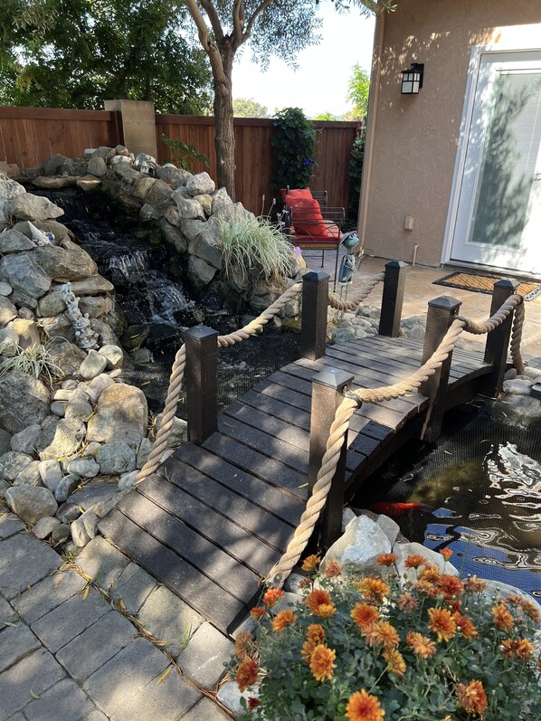 A wooden deck with a rope railing, a small pond with water lilies, and orange flowers in the foreground.