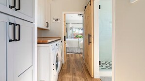 Un pasillo estrecho con una lavadora, una cama con cubierta blanca y una puerta corredera que da a una habitación con vistas.