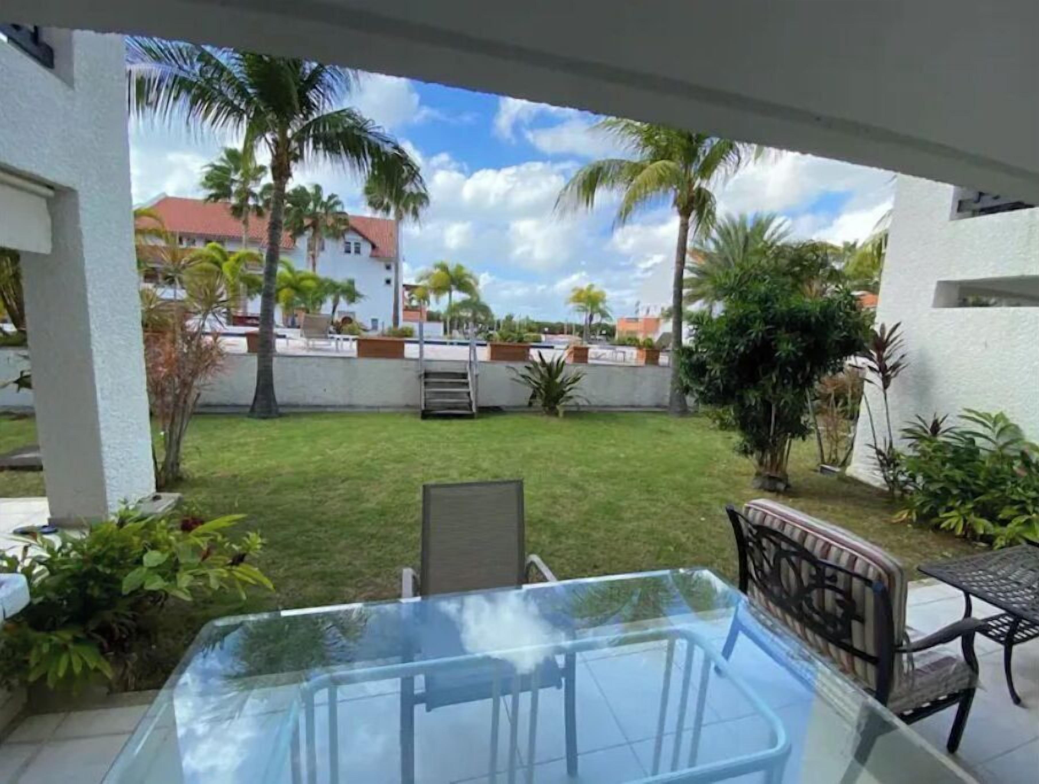 A glass-enclosed patio with a table and chairs, surrounded by a well-maintained lawn and palm trees.