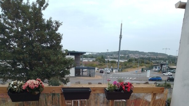 A balcony with a table and chairs, a view of a road and buildings, and hanging flower baskets.