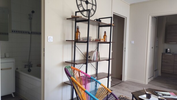 Een moderne woonkamer met een kleurrijke geweven stoel, een metalen boekenkast en een keuken op de achtergrond.