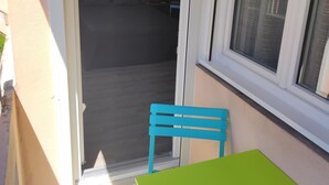 Een klein balkon met een groene tafel en een blauwe stoel, een raam met zonwering en uitzicht op een donkere kamer met een bed en een wandplank.