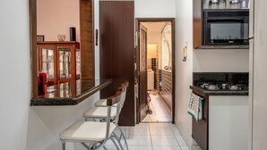 Une petite cuisine avec un plan de travail, un évier et une cuisinière. Une porte donne sur un couloir dont le sol est carrelé.