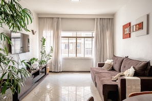 A modern living room with a large window, a brown sofa, a TV on a wooden stand, and several potted plants.