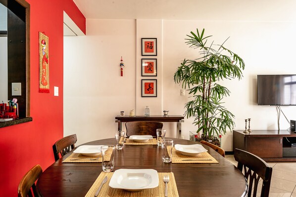 A dining area with a round table set for four, surrounded by wooden chairs. There is a large potted plant to the right and a flat-screen TV mounted on the wall.