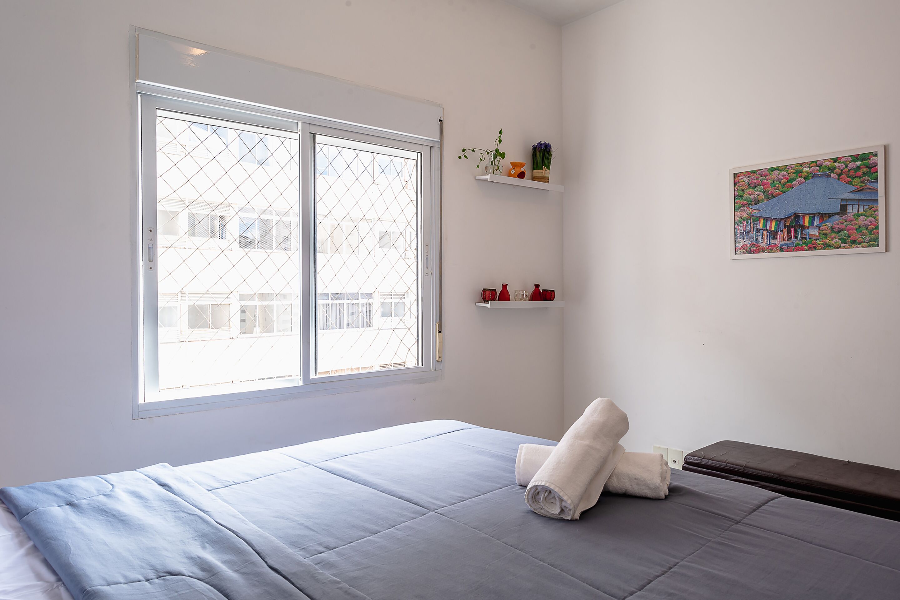 A room with a bed, a window, a painting, and a shelf with items.