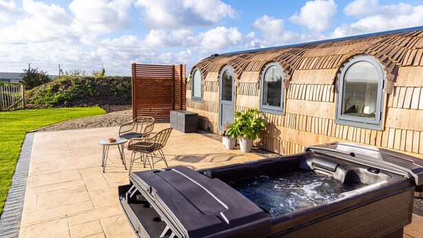 A hot tub with a wooden deck, a seating area with chairs, and a building with arched windows. - Indrek - Mayflower Meadow (Driffield)
