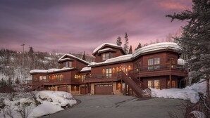 Una cabaña de troncos grande y de varios pisos, con techo cubierto de nieve, múltiples balcones y un telesilla al fondo.