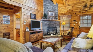 Un interior de cabaña acogedor, con paredes de madera, chimenea de piedra, televisor y asientos cómodos.