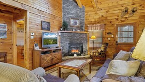 Acogedor interior de una cabaña de troncos con chimenea de piedra, muebles de madera y un televisor.