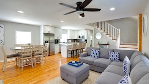 A modern living room with a grey sofa, ottoman, and wooden dining table with chairs. A kitchen with white cabinets and a microwave is visible in the background.