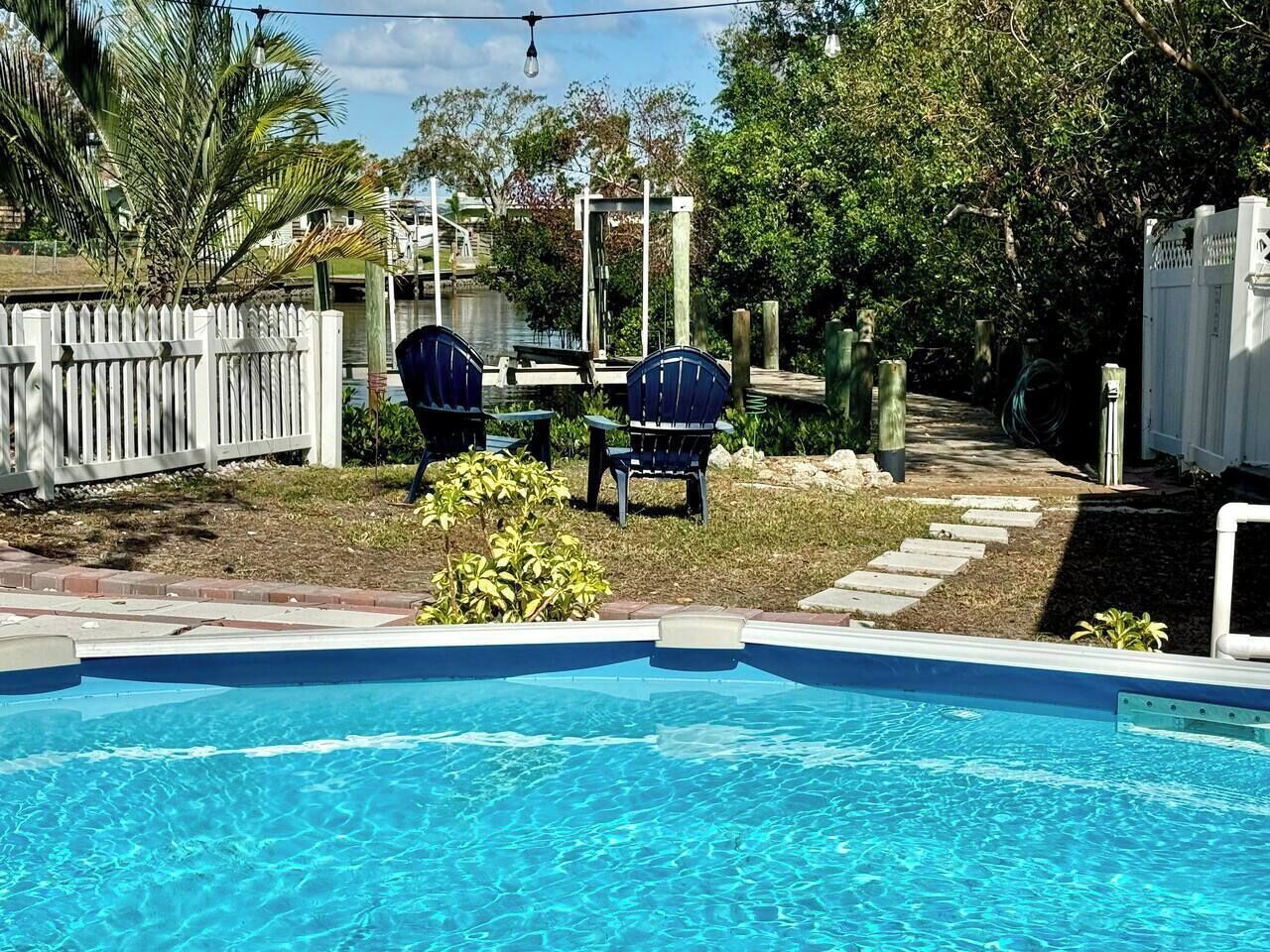 A swimming pool with two blue chairs and a table, surrounded by a white fence and greenery.