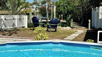 A swimming pool with two blue chairs and a table, surrounded by a white fence and greenery.