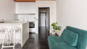 A modern kitchen with white cabinets, a marble countertop, and a built-in oven. There is a teal sofa and a potted plant in the living area. - Riverview Studio in Historical Heart of Antwerp (Antwerpen)