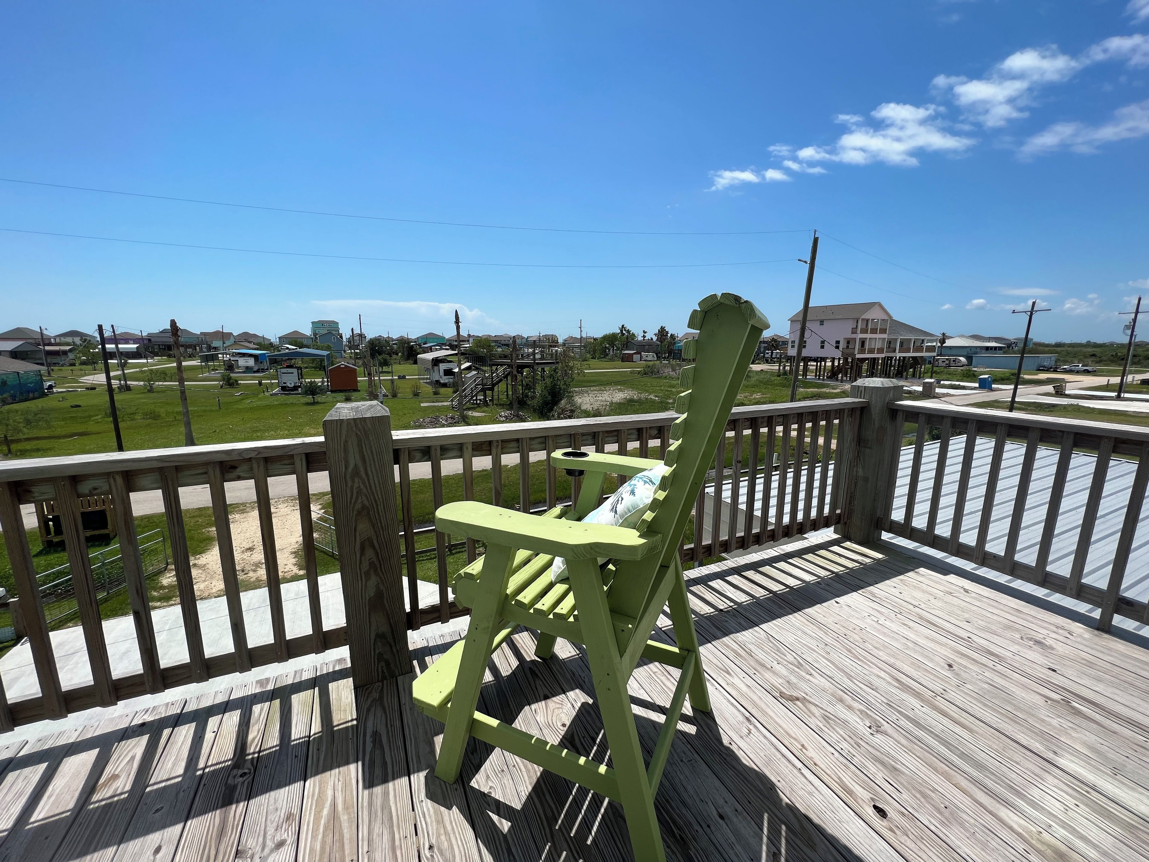 Een houten terras met een groene stoel, een leuning en uitzicht op een woonwijk met huizen en een weg.