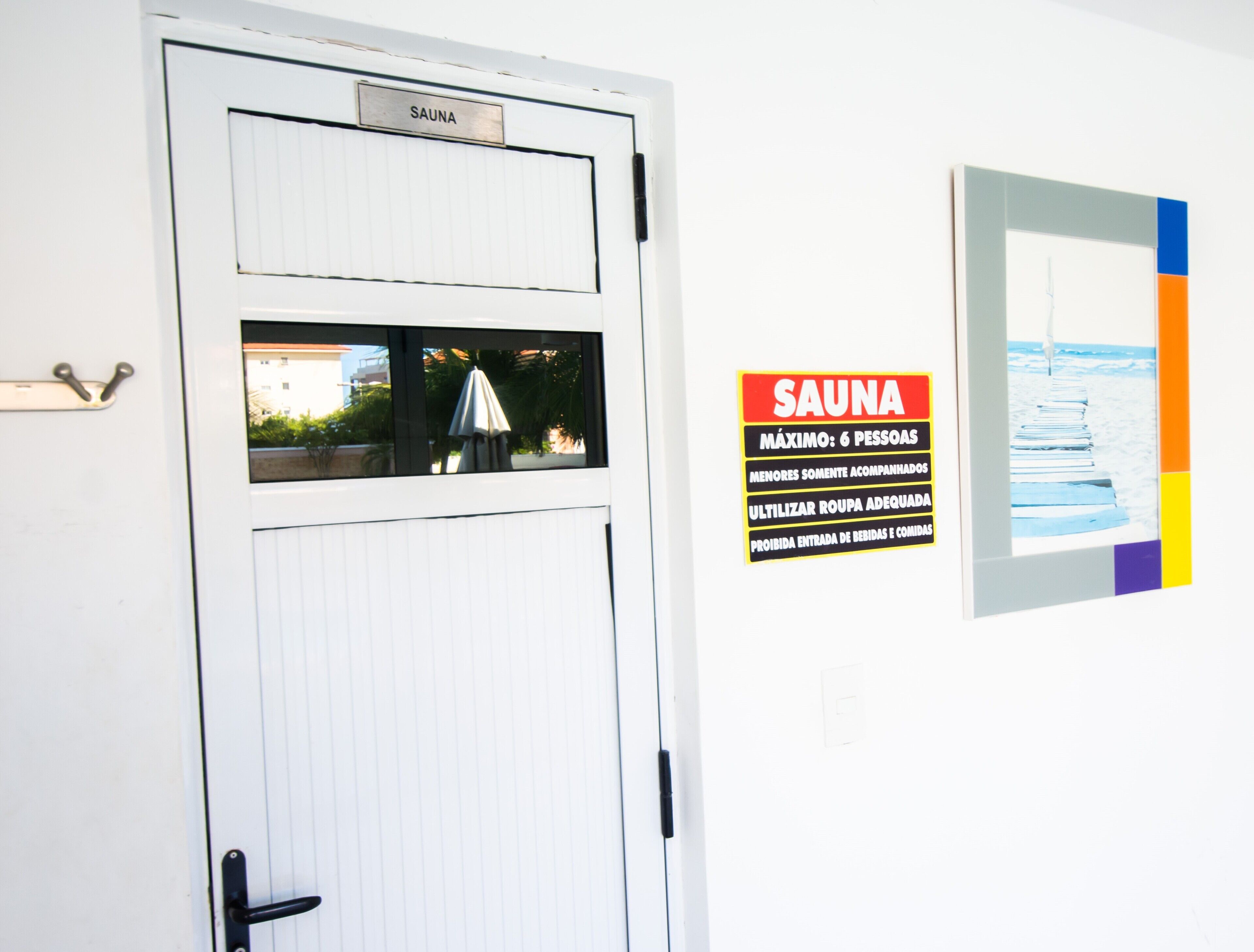 Eine Tür mit dem Schild 'SAUNA'. Auf dem Schild ist angegeben, dass maximal 6 Personen erlaubt sind, Speisen und Getränke nicht erlaubt sind, und es wird daran erinnert, geeignete Kleidung zu tragen.